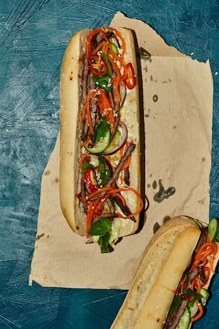 Vietnamese Banh Mi sandwich with grilled meat, pickled carrots, cucumber, and herbs on brown paper.